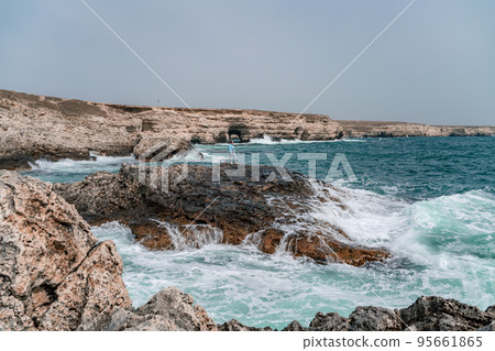 A woman in a blue jacket stands on a rock above a cliff above the sea and looks at the raging ocean. Girl traveler rests, thinks, dreams, enjoys nature. Peace and calm landscape, windy weather. 95661865