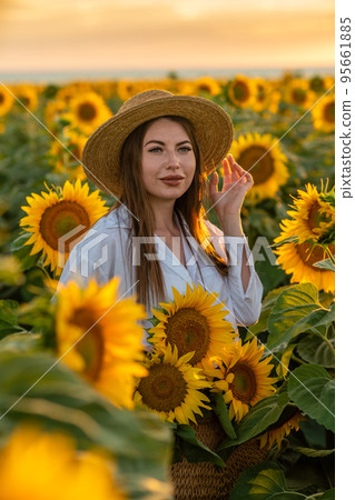 A girl in a hat on a beautiful field of sunflowers against the sky in the evening light of a summer sunset. Sunbeams through the flower field. Natural background. 95661885
