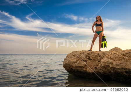 Young slim smiling woman standing on the rocky seashore with scuba diving equipment 95662020