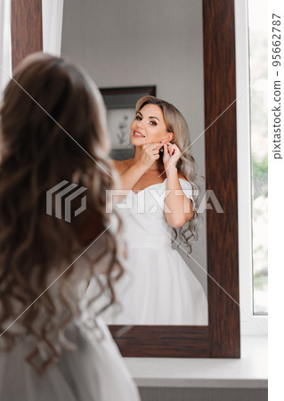 blonde bride in a white dress looks at the reflection and puts on earrings.  95662787