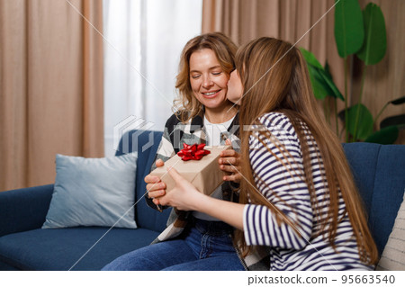 Adult daughter kissing happy mother who sitting on sofa and giving her present Adult daughter kissing happy mother who sitting on sofa and giving her present 95663540