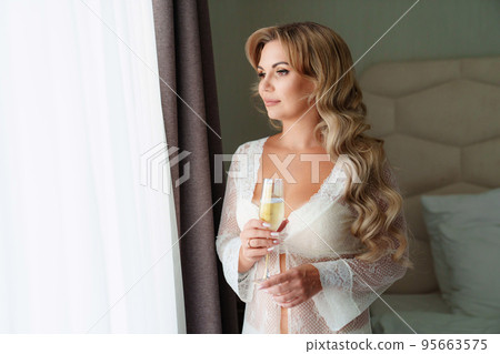 a blonde woman in a white peignoir with a glass of champagne by the window. 95663575