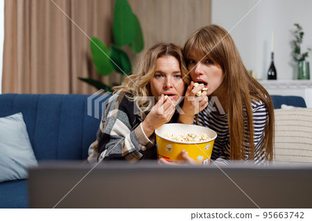 Two women of different ages watch horror movie and eat popcorn while sitting on the couch hugging each other 95663742
