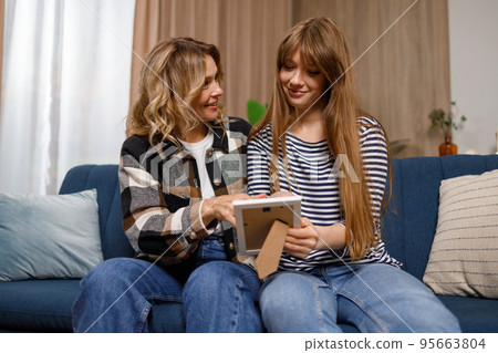 Mature woman and adult daughter smiling looking photo in frame at home Mature woman and adult daughter smiling looking photo in frame at home 95663804