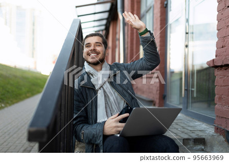 men sits with a laptop outside the office and greets a passing acquaintance with a gesture 95663969