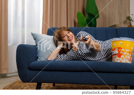 Cute young woman lying on cozy sofa in living room with remote control in hands, switching channels on TV 95664327