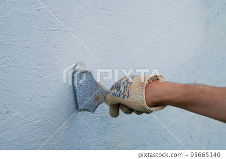 A hand in a work glove paints the wall of the house with blue paint. 95665140