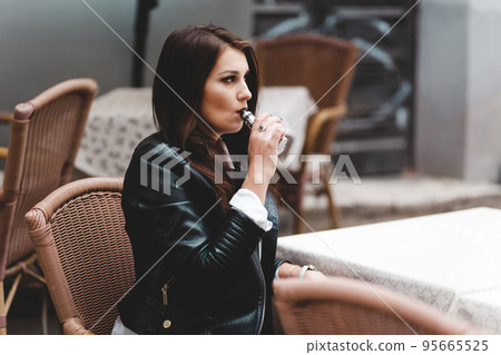 Stylish girl smokes a electronic cigarette. Serious woman with vape sitting on the table Stylish girl smokes a electronic cigarette. Serious woman with vape sitting on the table 95665525