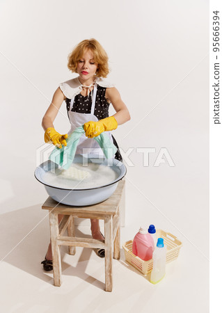 Portrait of stylish young woman in dress and apron doing washing isolated over white background. Housewife 95666394