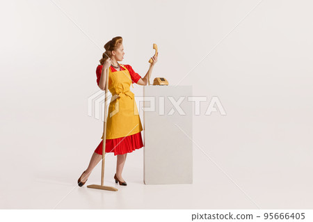 Portrait of elegant redhead woman with beautiful hairstyle cleaning with mop and talking on vintage phone isolated over white background 95666405