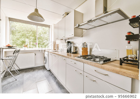 Fragment of interior of light narrow home kitchen with minimalist style white furniture with sink and stove Fragment of interior of light narrow home kitchen with minimalist style white furniture with sink and stove 95666902