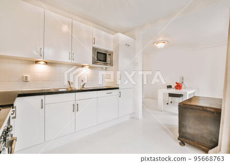 Fragment of interior of light narrow home kitchen with minimalist style white furniture with sink and stove Fragment of interior of light narrow home kitchen with minimalist style white furniture with sink and stove 95668763