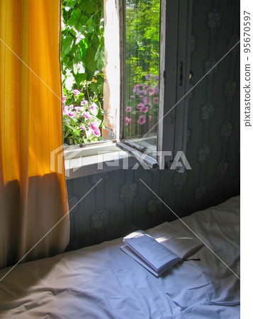 Summer day in the country. An open book lies on a bed near an open old window. Outside the window, petunia flowers bloom and grapes weave. Yellow curtains hang on the wall. Summer day in the country. An open book lies on a bed near an open old window. Outside the window, petunia flowers bloom and grapes weave. Yellow curtains hang on the wall. 95670597