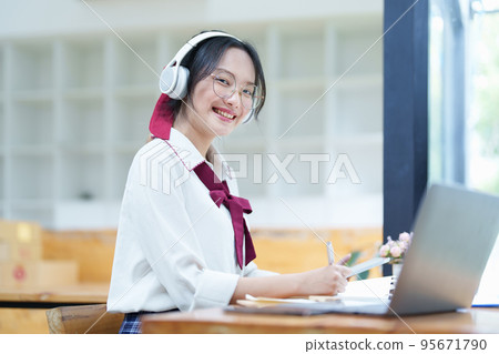 Portrait of a teenage Asian woman using a computer, wearing headphones and using a notebook to study online via video conferencing on a wooden desk in library. 95671790
