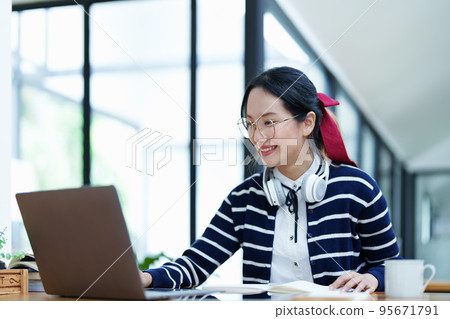 Portrait of a teenage Asian woman using a computer, wearing headphones and using a notebook to study online via video conferencing on a wooden desk in library. Portrait of a teenage Asian woman using a computer, wearing headphones and using a notebook to study online via video conferencing on a wooden desk in library. 95671791