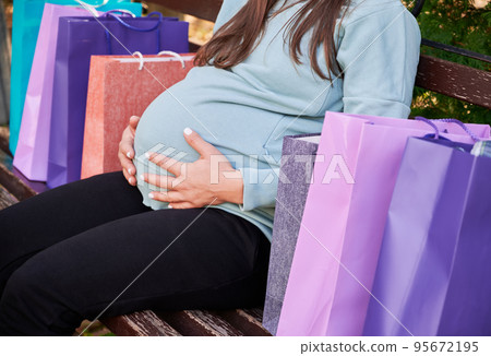 Cropped pregnant woman with colorful bags spending daytime in park after shopping of infant wear during expectation awaiting. Unrecognizable female with big belly abdomen resting at bench. Cropped pregnant woman with colorful bags spending daytime in park after shopping of infant wear during expectation awaiting. Unrecognizable female with big belly abdomen resting at bench. 95672195