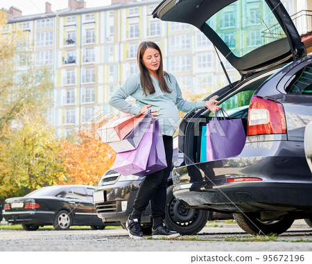 Woman with paper bags give child birth at auto parking in city downtown. Young woman dressed in casual wear feeling pain during broken water after shopping for awaiting baby 95672196