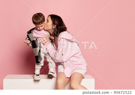 Portrait of young woman and little boy, mother kissing her son isolated on pink studio background. Mother's Day celebration. Concept of family, childhood, motherhood Portrait of young woman and little boy, mother kissing her son isolated on pink studio background. Mother's Day celebration. Concept of family, childhood, motherhood 95672522