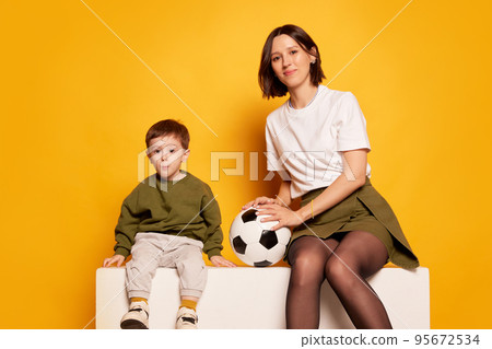 Charming young woman with kid, mother and her little son holding football ball isolated on bright yellow background. Sport, hobbies, games, Mother's day concept Charming young woman with kid, mother and her little son holding football ball isolated on bright yellow background. Sport, hobbies, games, Mother's day concept 95672534
