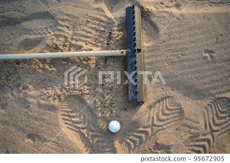 Golf concept. Golf ball and rake sand on bunker in beautiful golf course at sunset background Golf concept. Golf ball and rake sand on bunker in beautiful golf course at sunset background 95672905