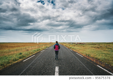 Female tourist running on straight road through farmland and stormy sky in countryside on summer 95675378