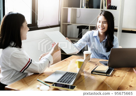 Two female company employees happily smiled at each other as they handed each other folders Two female company employees happily smiled at each other as they handed each other folders 95675665
