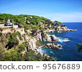 Panoramic photo of the rocky seashore with the sky, with green plants, trees and private houses. The Spanish coast of the Costa Brava, the Mediterranean Sea. Tourism, living in a beautiful place. 95676889