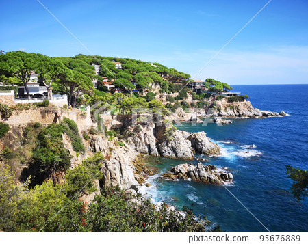 Panoramic photo of the rocky seashore with the sky, with green plants, trees and private houses. The Spanish coast of the Costa Brava, the Mediterranean Sea. Tourism, living in a beautiful place. Panoramic photo of the rocky seashore with the sky, with green plants, trees and private houses. The Spanish coast of the Costa Brava, the Mediterranean Sea. Tourism, living in a beautiful place. 95676889
