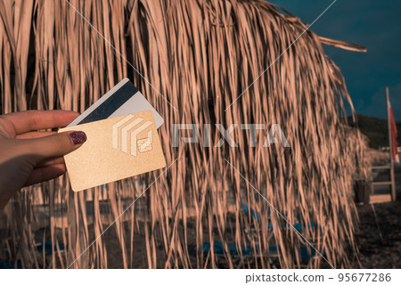 Golden and White Bank Card In Woman Hand On Background Of Beach Umbrellas Made Of Palm Leaves In Moraitika, Corfu, Greece. The Concept Of Payment For Relax And Unlimited Possibilities. Copy Space. Golden and White Bank Card In Woman Hand On Background Of Beach Umbrellas Made Of Palm Leaves In Moraitika, Corfu, Greece. The Concept Of Payment For Relax And Unlimited Possibilities. Copy Space. 95677286