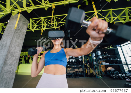 Woman wearing glasses of the virtual reality exercising with dumbbells Woman wearing glasses of the virtual reality exercising with dumbbells 95677487