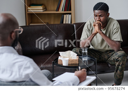African young man talking about his problems to psychology while sitting on sofa at his office African young man talking about his problems to psychology while sitting on sofa at his office 95678274
