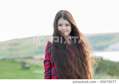 Little girl with long brown hair in red tartan dress standing on the grass, nature on the background, Cuckmere beach in East Sussex, UK 95679077