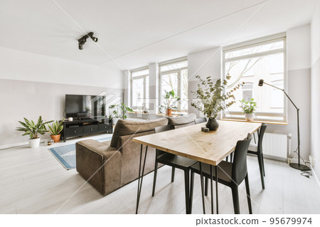 Simple interior of light living room with gray sofa in front of TV and dining table against window Simple interior of light living room with gray sofa in front of TV and dining table against window 95679974