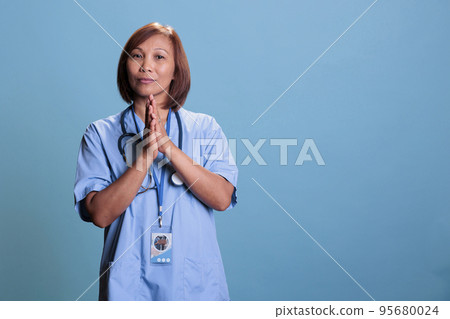 Religious medical assistant holding hands praying to god during patient checkup visit. Elderly asian nurse wearing medical blue uniform and stethoscope, health care service concept Religious medical assistant holding hands praying to god during patient checkup visit. Elderly asian nurse wearing medical blue uniform and stethoscope, health care service concept 95680024