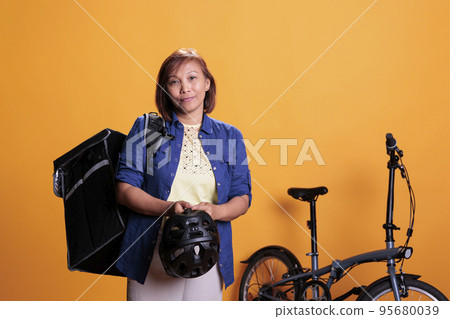 Elderly deliverywoman carrying thermal delivery backpack standing beside bike in studio with yellow background. Restaurant worker delivering pizza for lunch. Food transportation service and concept 95680039