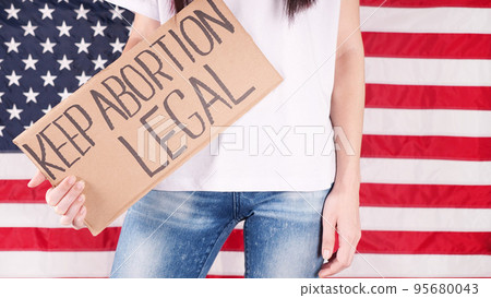 Young woman protester holds cardboard with Keep Abortion Legal sign against USA flag on background. Girl protesting against anti-abortion laws. Feminist power. Equal opportunity Womens rights reedom. 95680043
