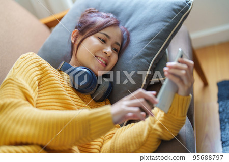 Portrait of a smiling Asian woman wearing a pair of headphones and using her phone and listening to music while sitting on the sofa. Portrait of a smiling Asian woman wearing a pair of headphones and using her phone and listening to music while sitting on the sofa. 95688797