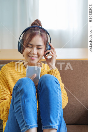 Portrait of a smiling Asian woman wearing a pair of headphones and using her phone and listening to music while sitting on the sofa. 95688808