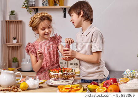 Little friends are making a cake together at a kitchen against a white wall with shelves on it. 95688869