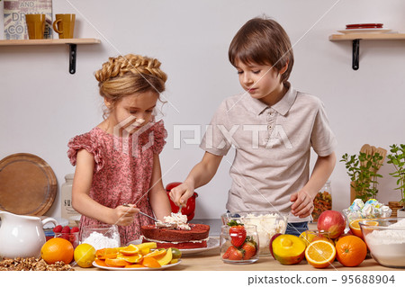 Little friends are making a cake together at a kitchen against a white wall with shelves on it. Little friends are making a cake together at a kitchen against a white wall with shelves on it. 95688904