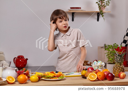 Cute little boy with brown hair is cooking at a kitchen against a white wall with shelves on it. Cute little boy with brown hair is cooking at a kitchen against a white wall with shelves on it. 95688978