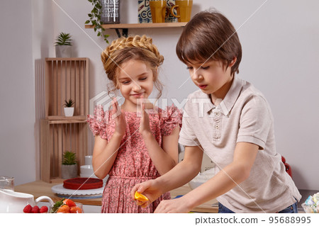 Cute kids are cooking together in a kitchen against a white wall with shelves on it. 95688995