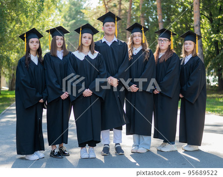 Seven graduates in robes stand in a row outdoors. 95689522