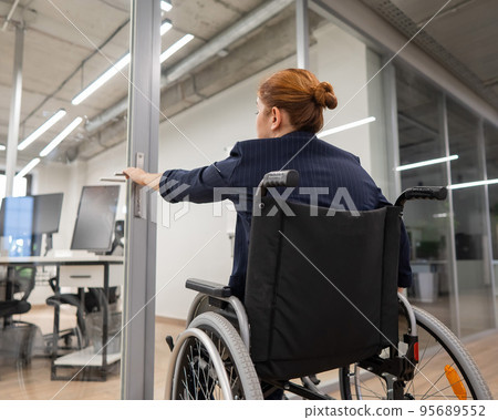 Red-haired caucasian woman in a wheelchair trying to open the door in the office. Red-haired caucasian woman in a wheelchair trying to open the door in the office. 95689552