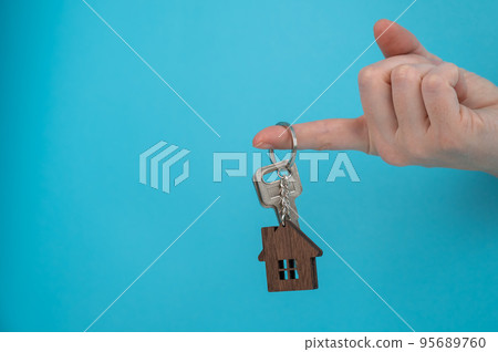 A woman's hand holds the keys with a keychain in the shape of a house on a blue background.  95689760
