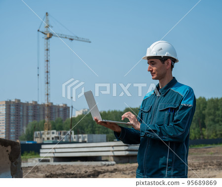 A male builder in a helmet stands against the background of a construction site with a laptop in his hands. A male builder in a helmet stands against the background of a construction site with a laptop in his hands. 95689869