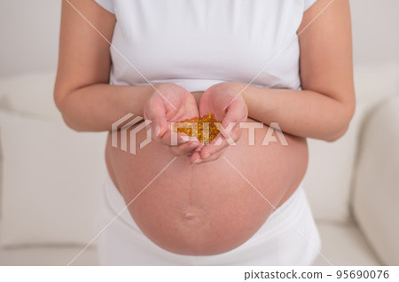 A pregnant woman with a bare belly holds a handful of vitamins. Transparent golden pills. Vitamin D for the expectant mother. A pregnant woman with a bare belly holds a handful of vitamins. Transparent golden pills. Vitamin D for the expectant mother. 95690076