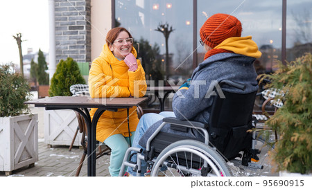 Two girlfriends in a cafe on a street terrace in winter. Woman in a wheelchair. 95690915