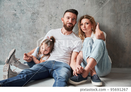 Loving parents and little child sitting by the wall in new apartment. Woman and man holding hands and looking at camera while cuddling with daughter in new home. 95691267