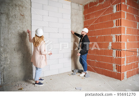 Full length of woman standing by brick wall and discussing apartment replanning and renovation with specialist. Female homeowner having meeting with builder in building under construction. 95691270